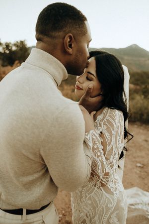 Groom tenderly kisses bride's forehead in desert landscape as she wears intricate long-sleeve lace gown.