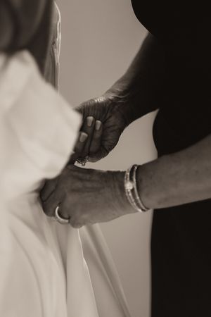 Black and white close-up of elder hands carefully fastening buttons on back of bride's white wedding dress.