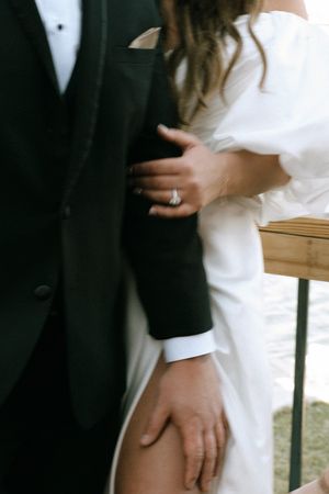 Bride in white off-shoulder gown holds grooms black tuxedo arm, showcasing diamond engagement ring.