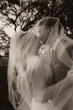 Bride and groom kissing under a flowing veil outdoors in black and white.