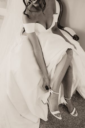 Black and white photo of smiling bride in off-shoulder gown with pearl ankle strap heels seated in chair.