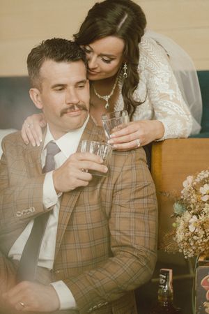 Groom in plaid brown suit holds whiskey glass as bride in lace wedding dress embraces him from behind.