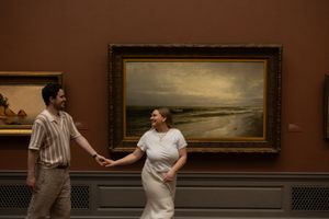 Couple holding hands and smiling in front of a landscape painting in a museum.