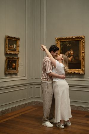 Couple sharing a romantic kiss in an art museum gallery surrounded by ornate gold-framed paintings.