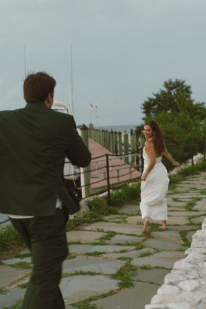 Bride in white dress smiling back at groom along a scenic waterfront pathway.