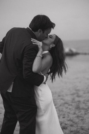 Black and white beach wedding photo of bride and groom sharing a passionate kiss at sunset.