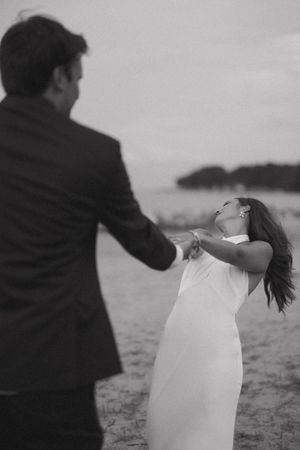 Black and white photo of bride in white gown laughing joyfully while holding grooms hands on beach.