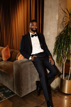 Elegant groom in black tuxedo with bow tie sits smiling on velvet sofa in warmly lit venue.