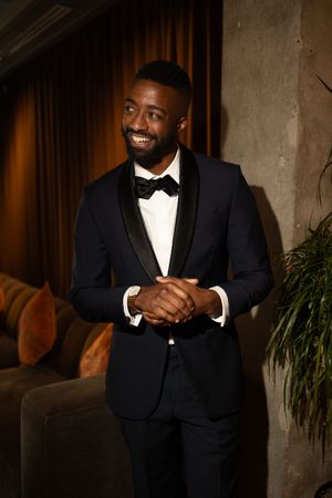 Handsome groom in black tuxedo with bow tie smiles confidently in warmly lit wedding venue setting.