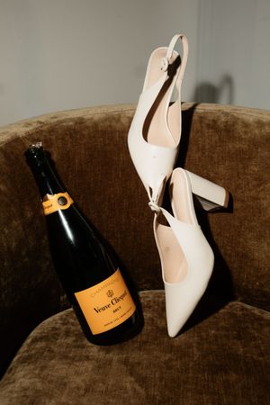 Veuve Clicquot champagne bottle beside white slingback heels on velvet chair, elegant wedding detail.