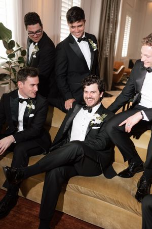 Groom laughing with groomsmen all dressed in black tuxedos relaxing on a sofa before the wedding.