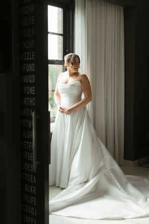Bride in a strapless white satin ball gown standing by a window in soft natural light.