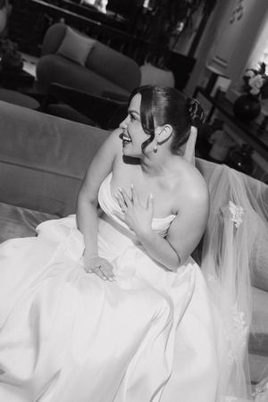 Smiling bride in strapless white ball gown sits on sofa in black and white wedding portrait photo.