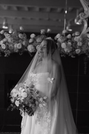 Elegant bride in strapless gown and cathedral veil holding lush bouquet before floral arrangement.