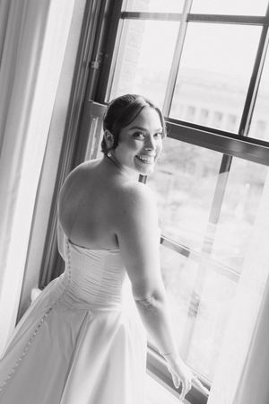 Black and white portrait of smiling bride in strapless gown looking back over shoulder at window.