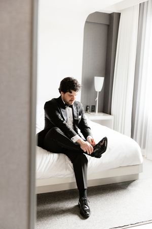 Groom in a black tuxedo sitting on a hotel bed putting on his dress shoes before the wedding.