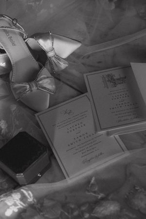 Black and white flat lay of bridal heels, wedding rings, and elegant wedding invitations on tulle.