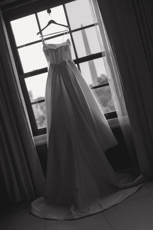 Black and white photo of a strapless ball gown wedding dress hanging in a window on a hanger.