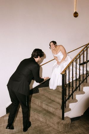 Bride in white gown descends elegant staircase as groom in black suit reaches up to greet her at wedding venue.