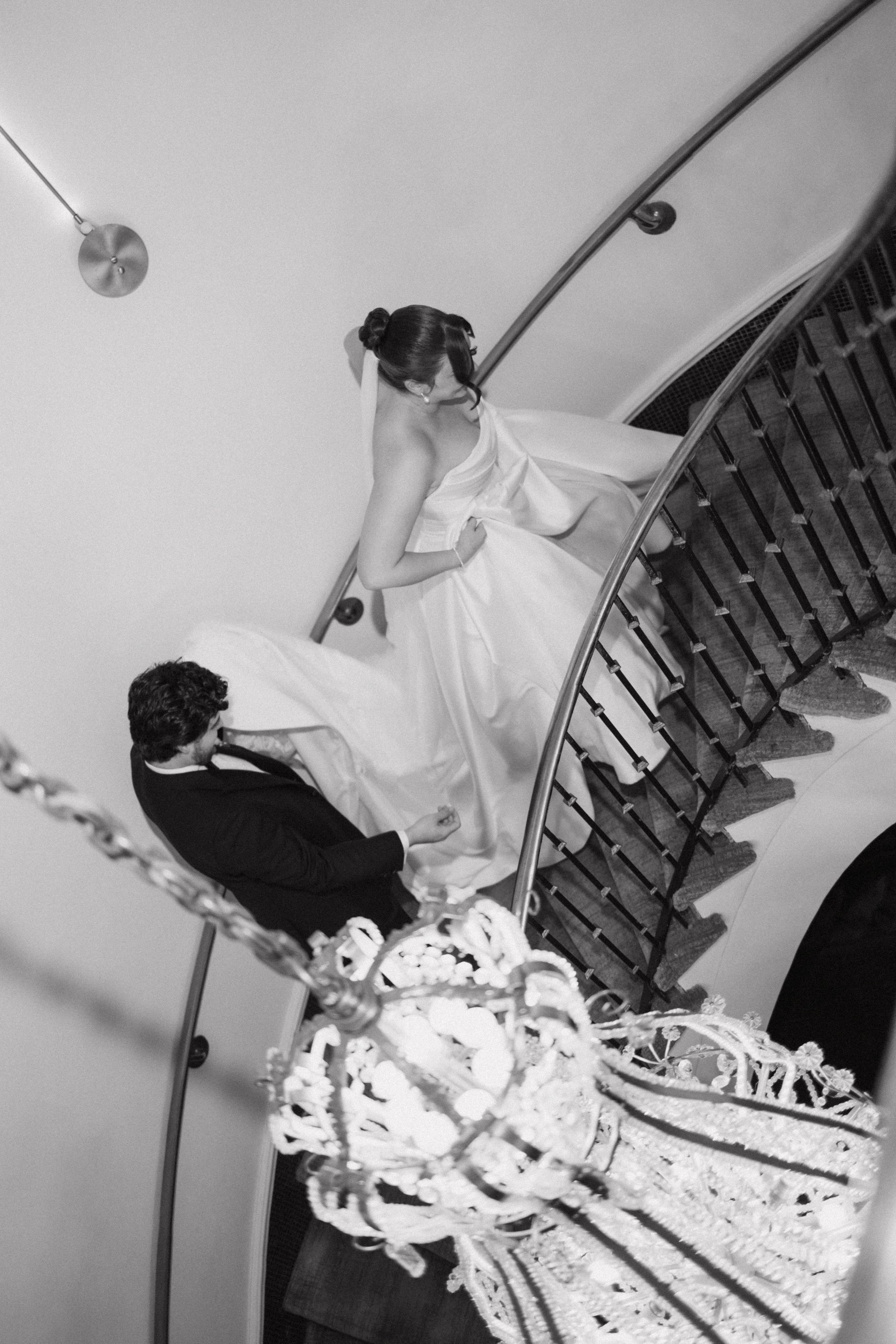 Aerial black and white view of bride and groom ascending curved staircase with chandelier below.