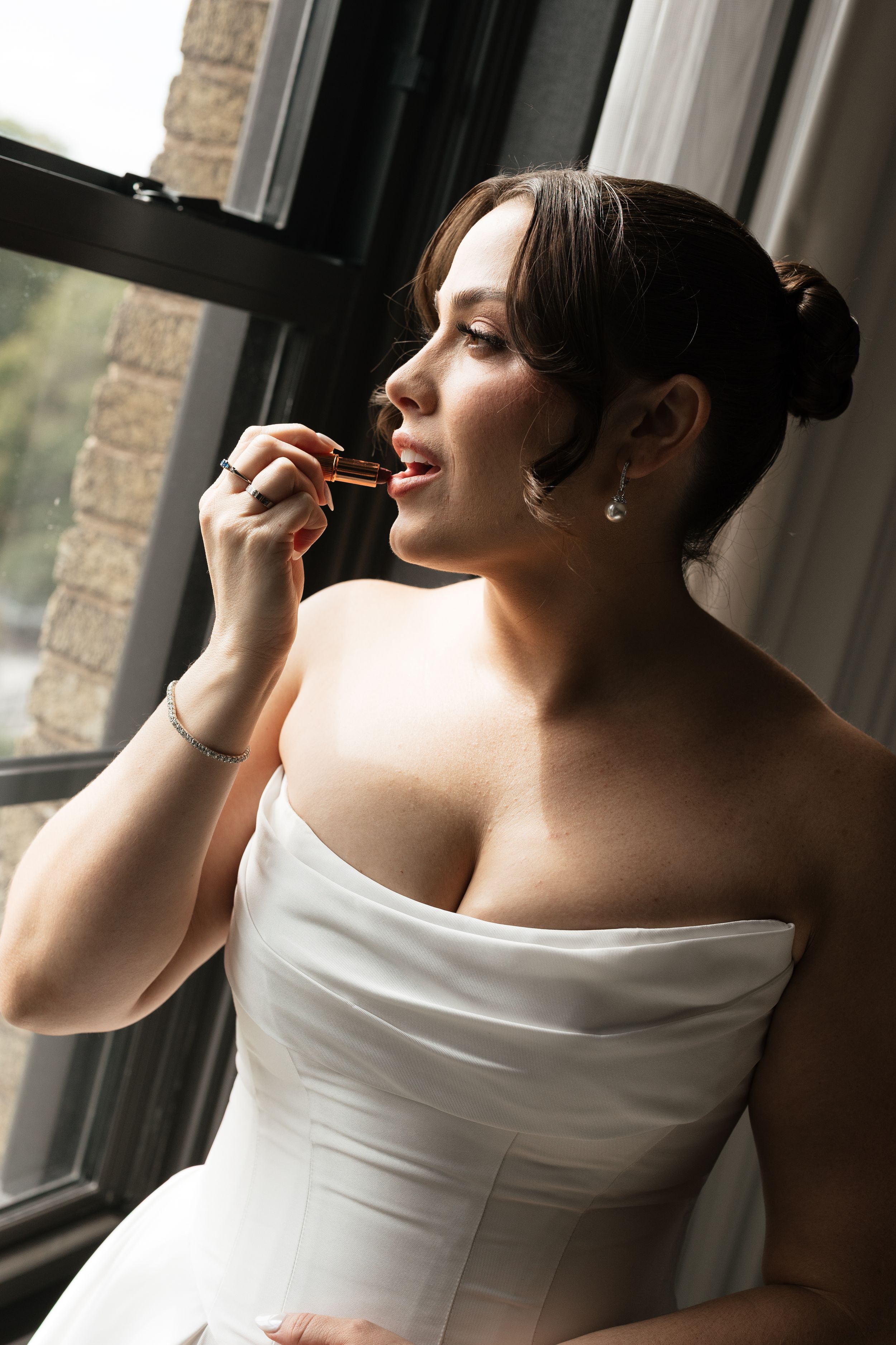 Elegant bride in white strapless wedding dress applying lipstick by a sunlit window.