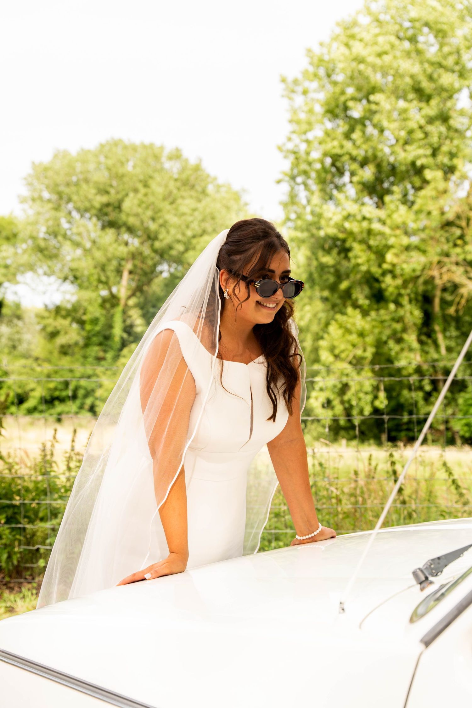 Person in a wedding dress and sunglasses leans against a white classic car in a sunny garden setting.