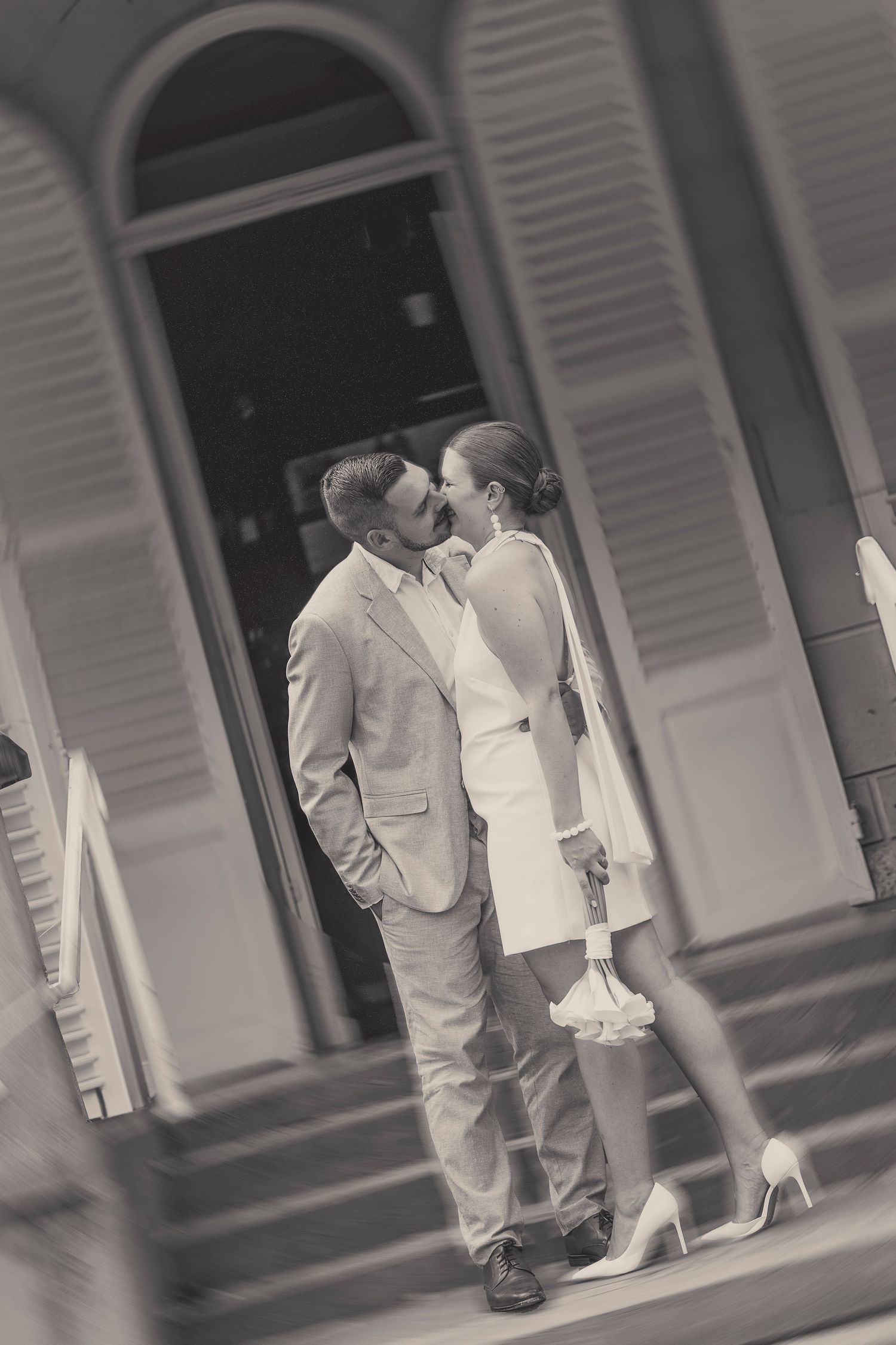 Black and white photo of couple in formal wear embracing by shuttered windows.