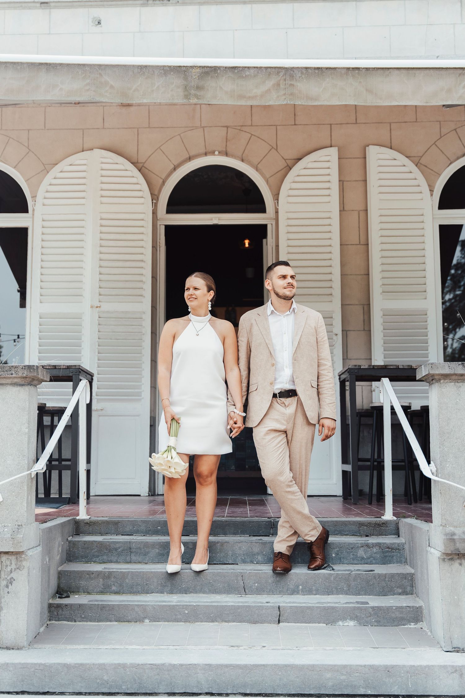 A stylish couple in formal wear standing on steps of a historic building with white shutters.