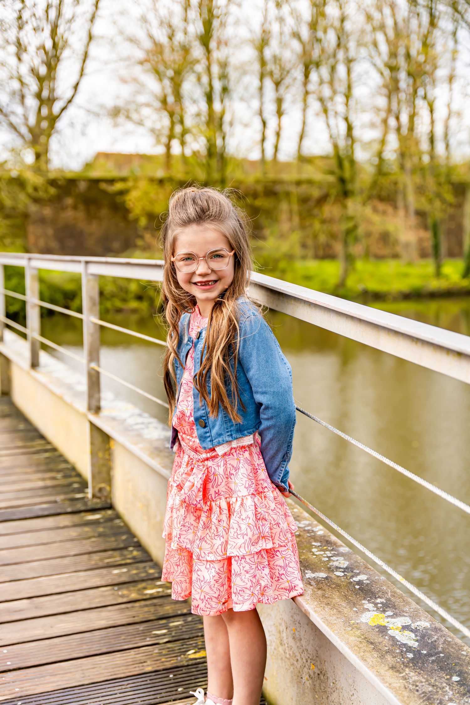 Child in blue denim jacket and pink dress posing on wooden bridge over water.