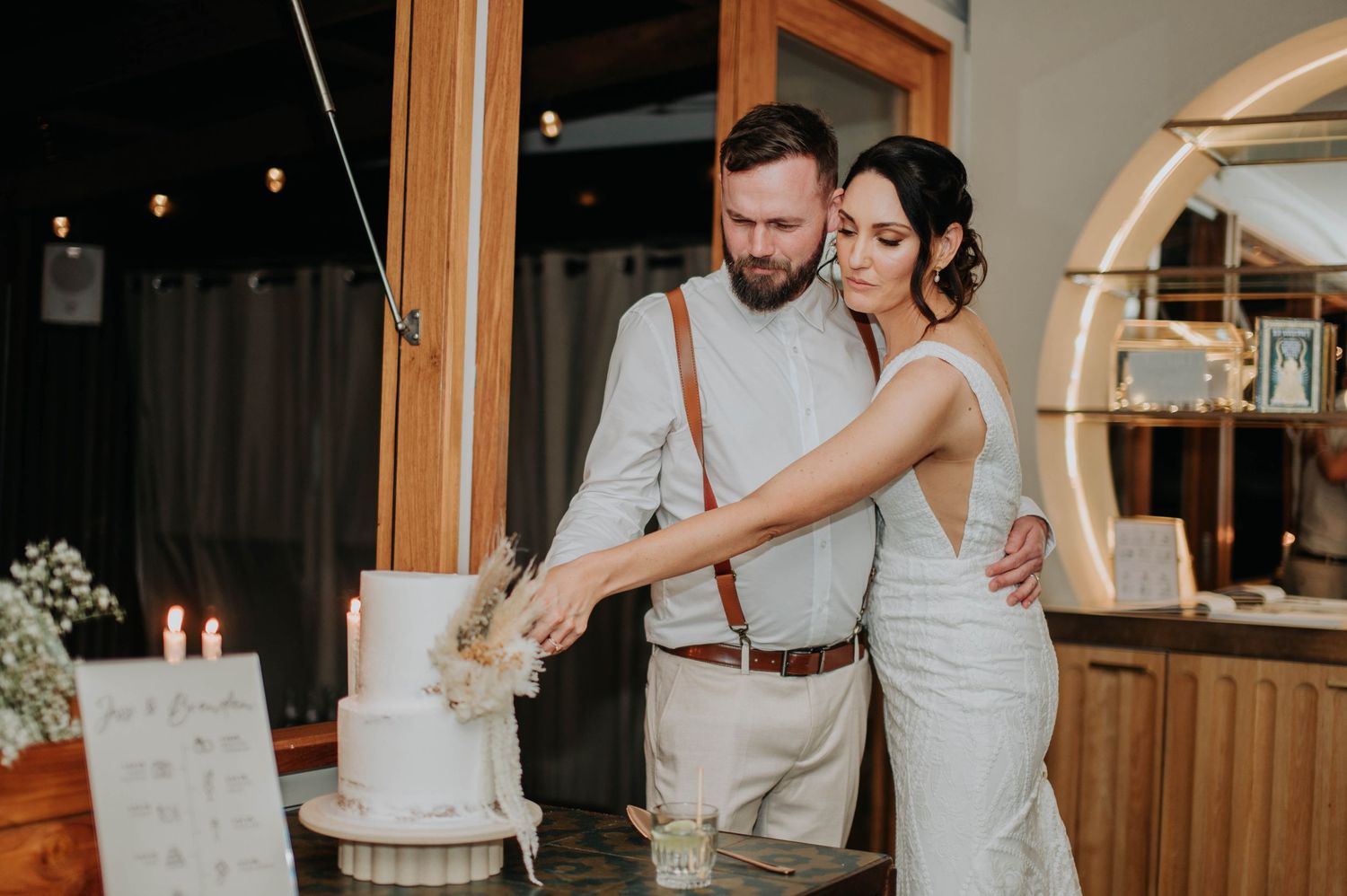 Couple in wedding attire cutting their white tiered wedding cake together indoors.