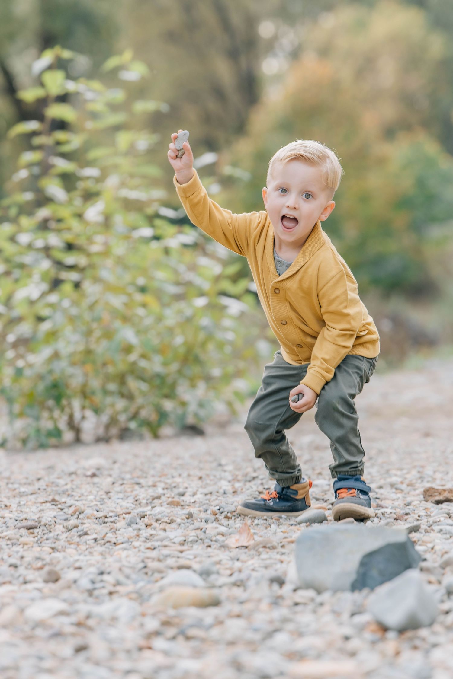 Energetic toddler boy in yellow cardigan throwing rocks on a pebble beach outdoors.