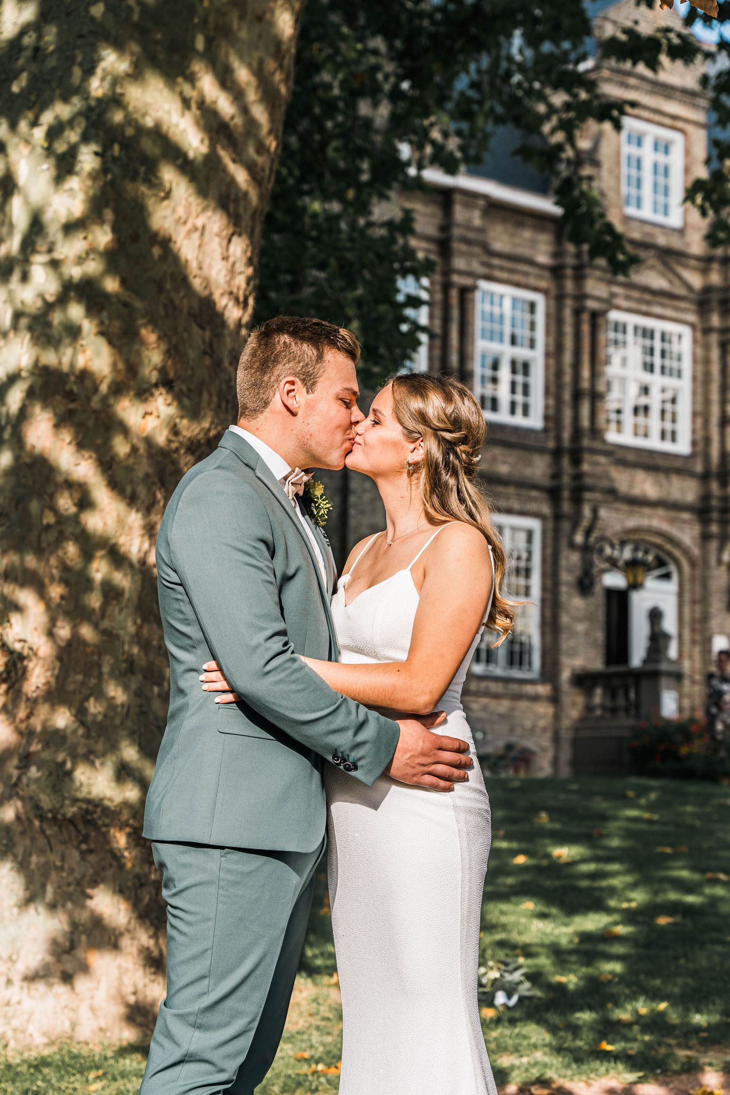 Romantic wedding moment in front of historic brick building with dappled sunlight.
