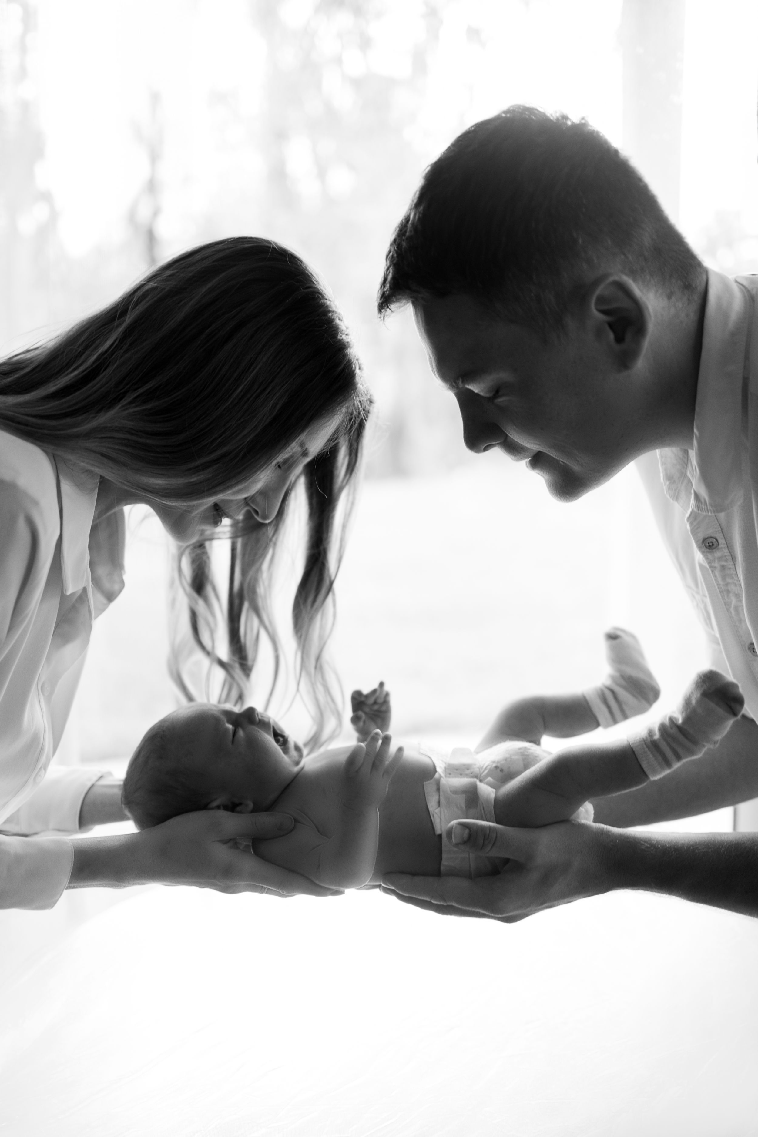 A tender black and white portrait of parents bonding with their newborn baby on a soft white surface.