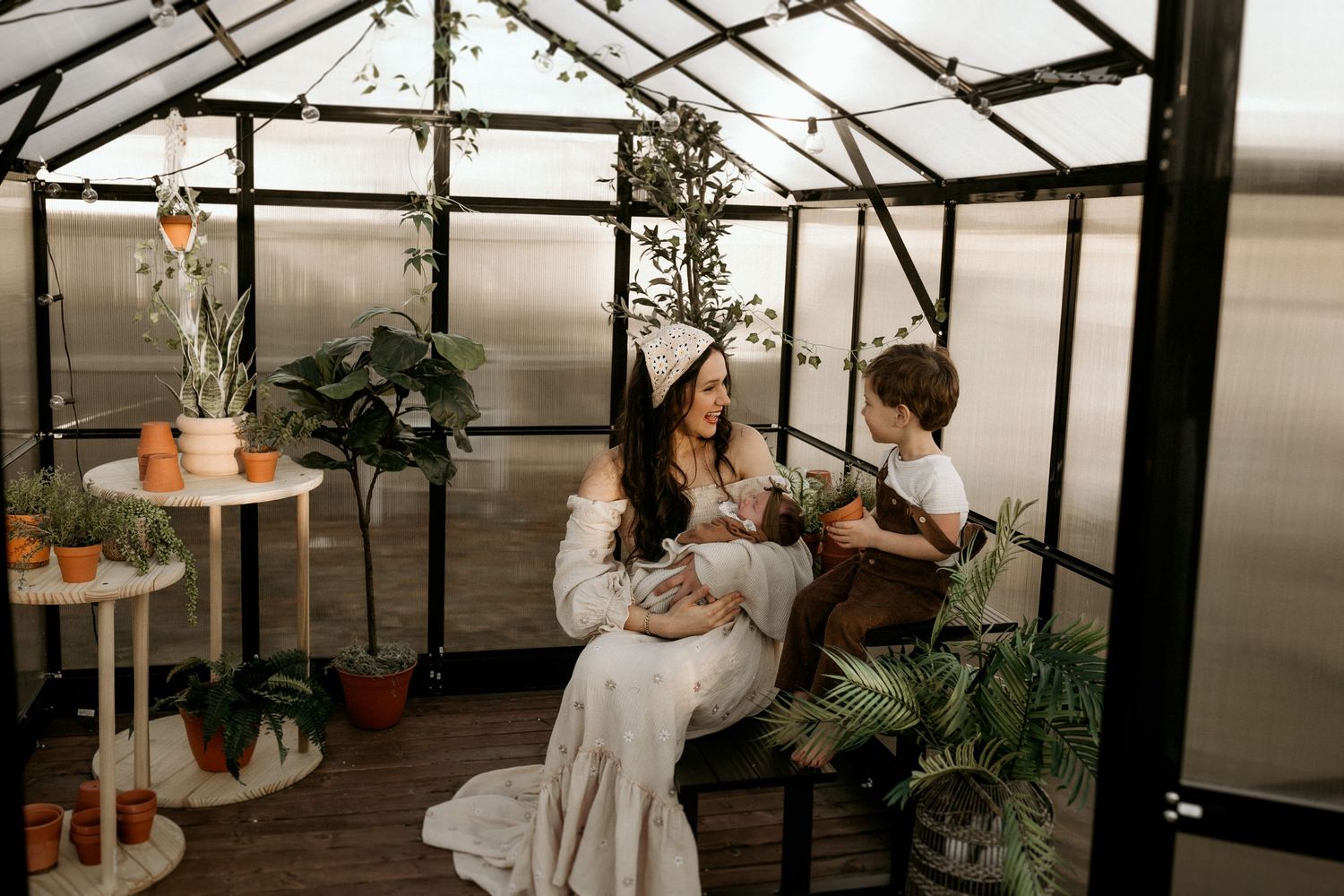 Family relax together in a cozy greenhouse setting with potted plants and natural light streaming through glass panels.