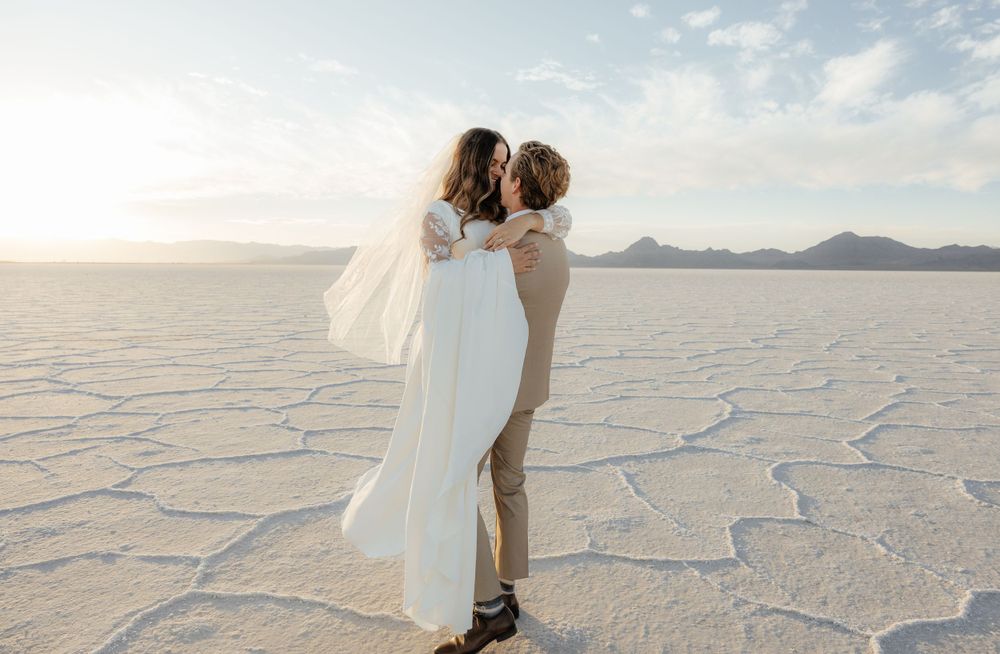 Groom lifts bride on the Bonneville Salt Flats with mountains and soft sunset sky behind them.