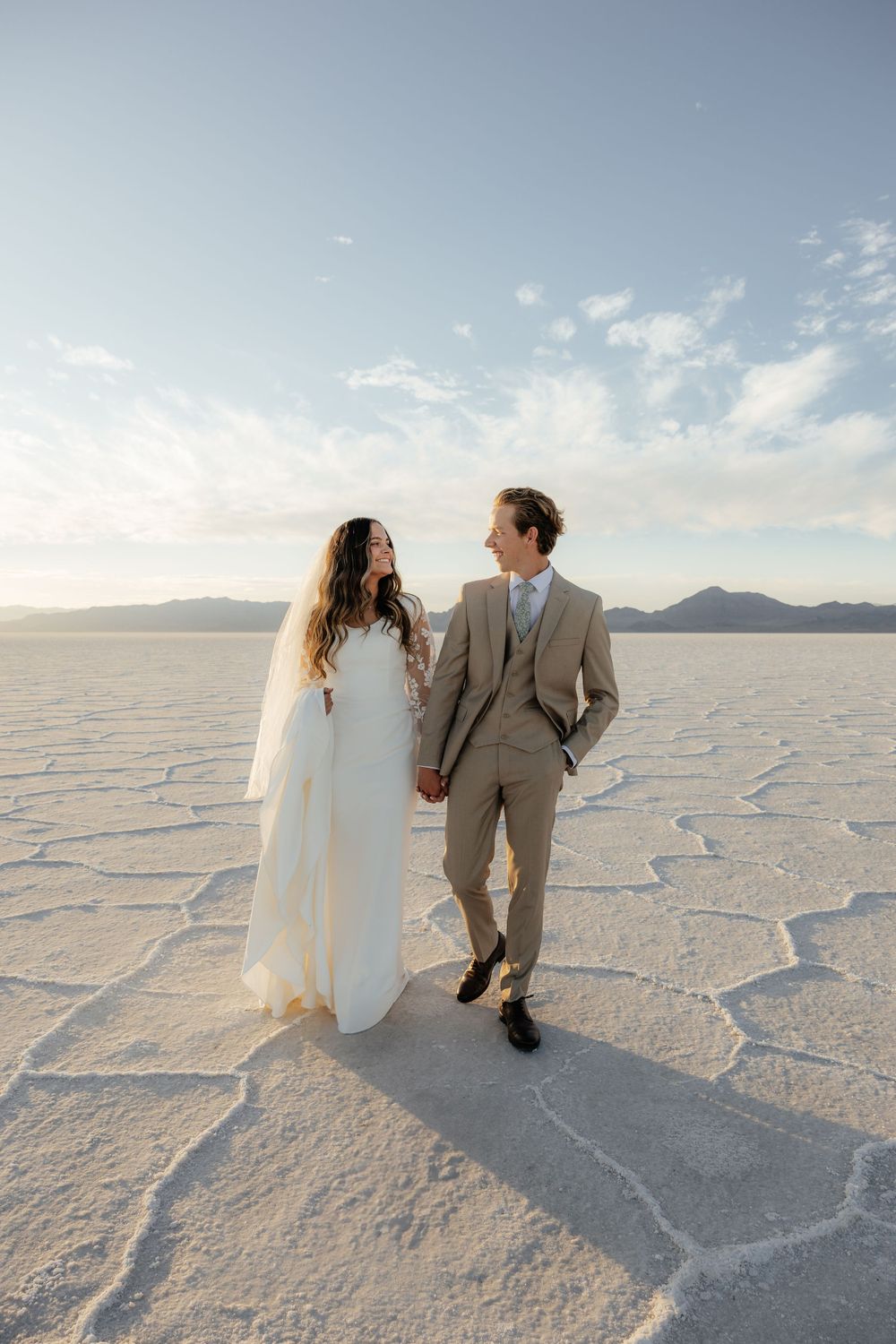 Bride and groom walk hand in hand smiling at each other on the vast white Bonneville Salt Flats.