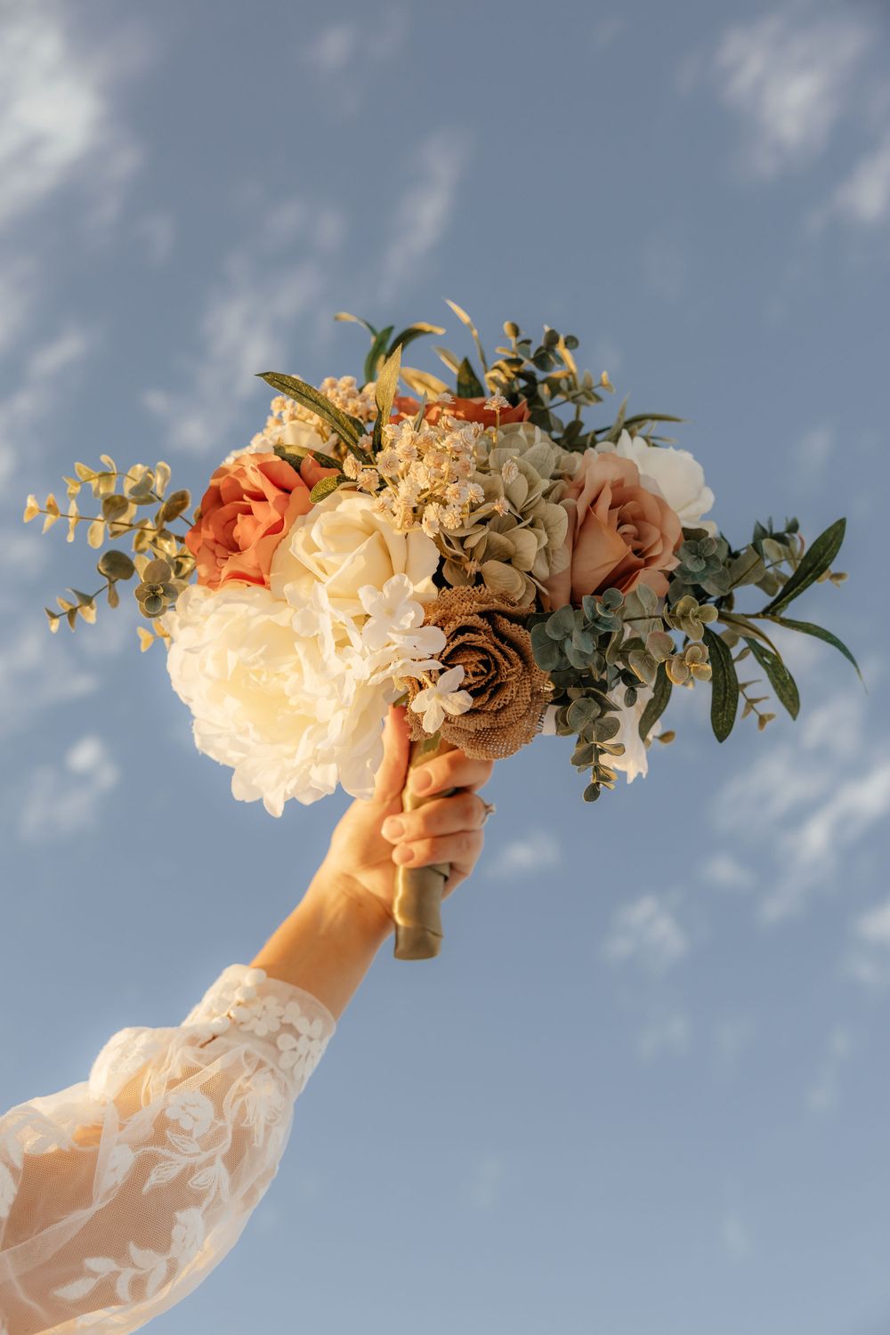 Bride holds a stunning wedding bouquet of peonies, roses, and eucalyptus raised against a blue sky.
