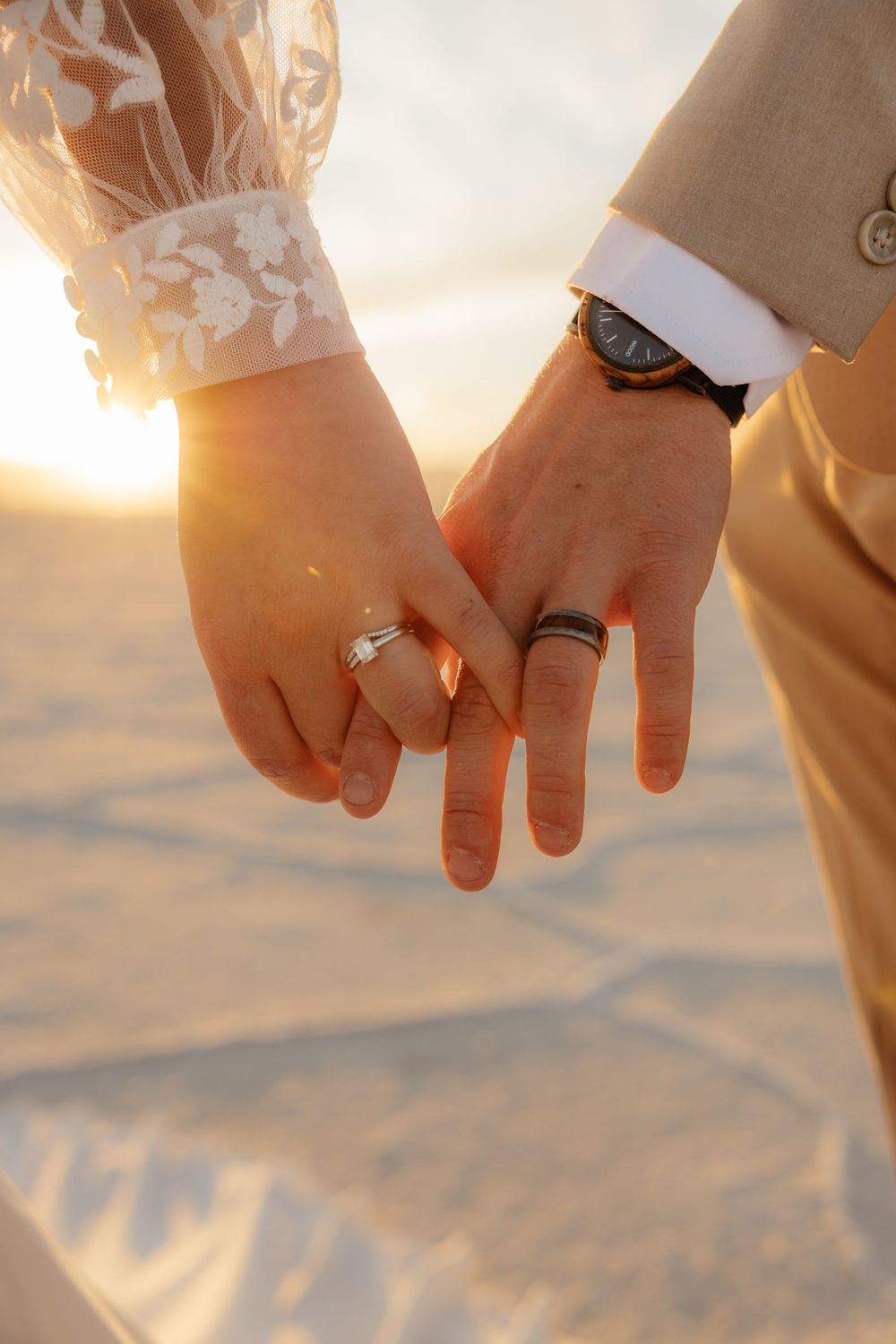 Close-up of bride and groom holding hands showing wedding rings at golden sunset on the salt flats.