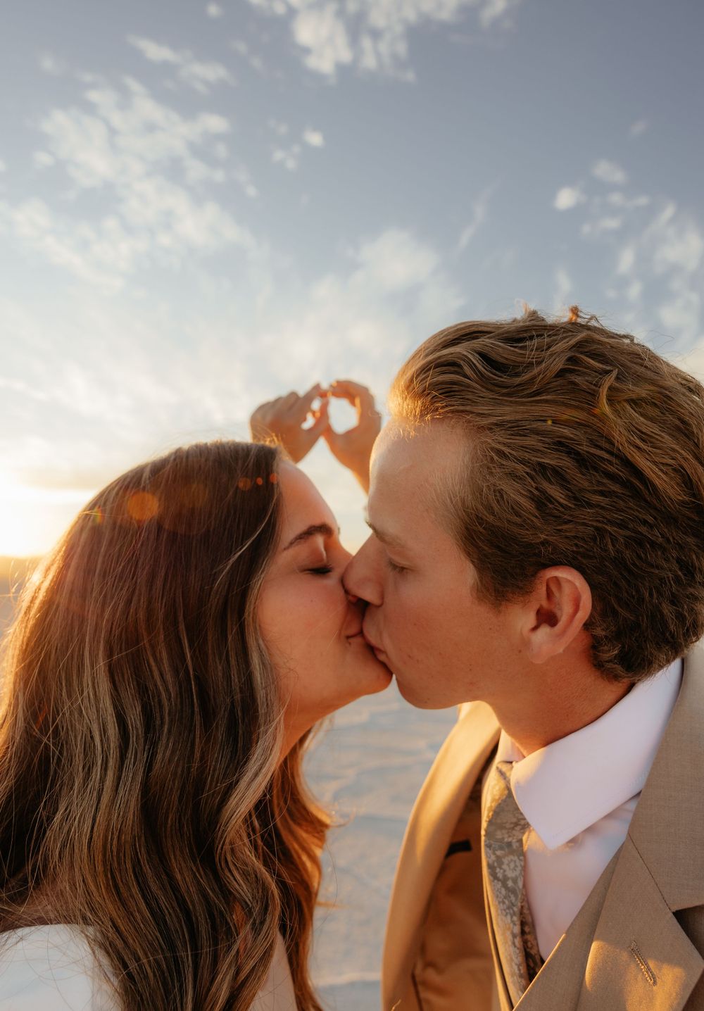 Couple shares a romantic kiss at golden sunset on the salt flats while forming a heart with their hands.