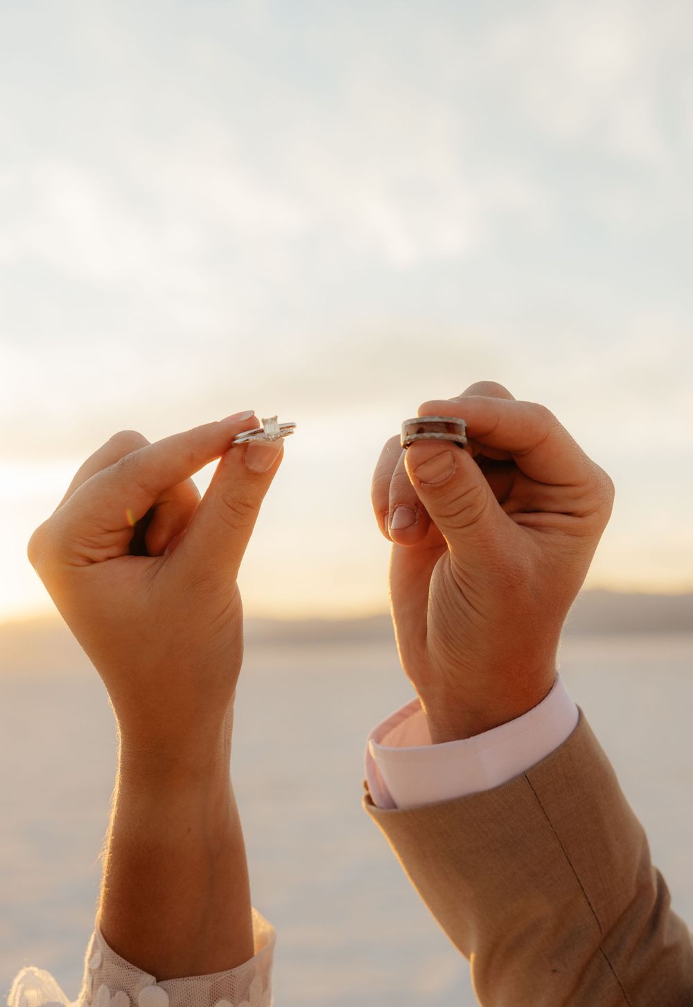 Bride and groom hold their wedding rings up against a soft pastel sunset sky on the salt flats.