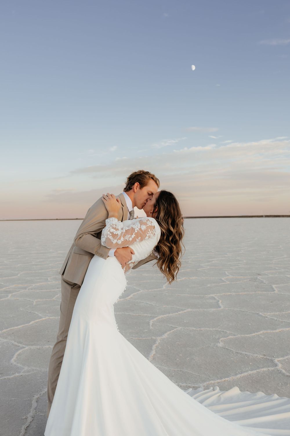 Bride and groom share a romantic kiss on the Bonneville Salt Flats at sunset with the moon visible above.