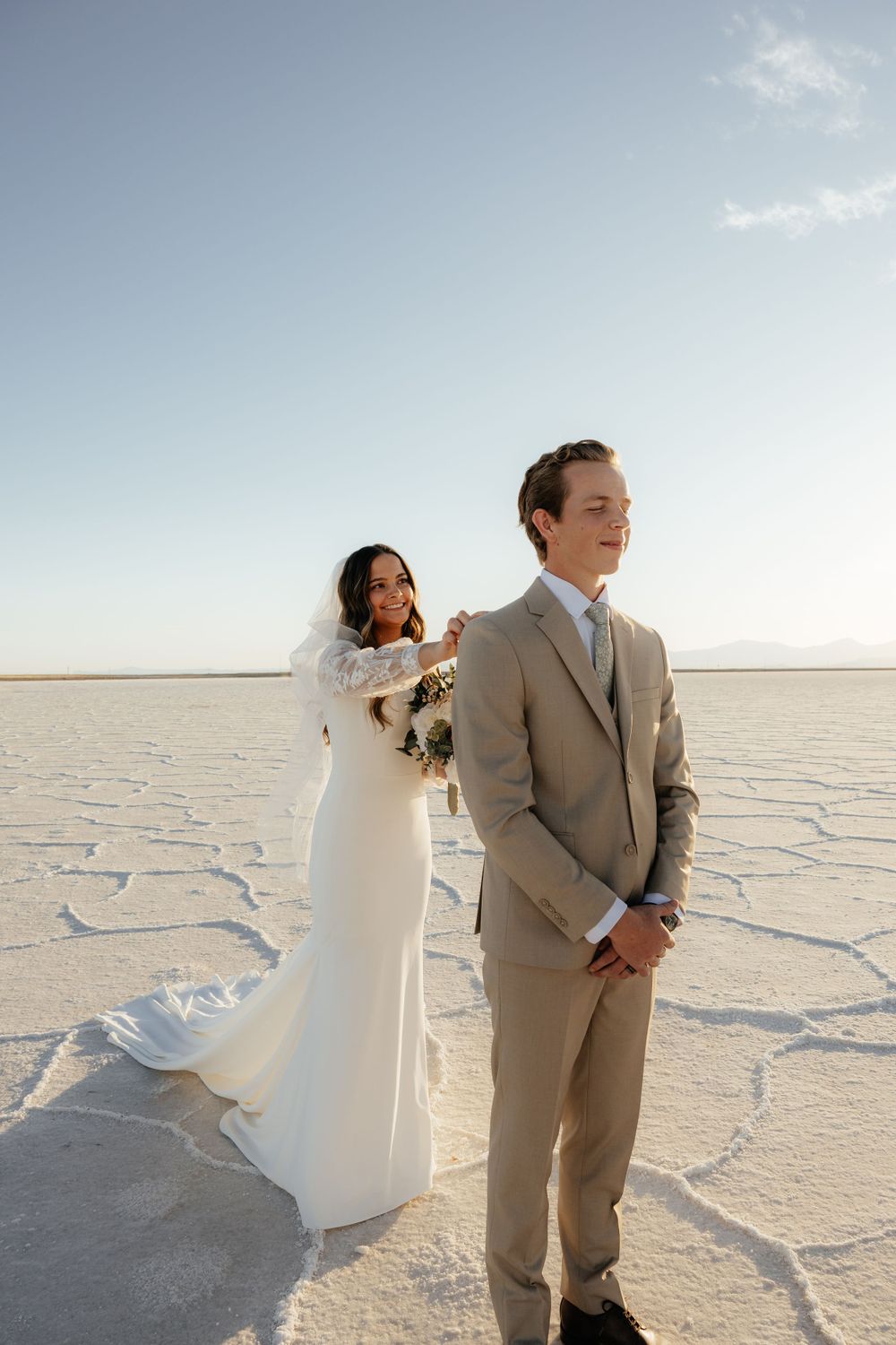 Bride in lace gown adjusts groom's suit jacket during first look on the Bonneville Salt Flats.