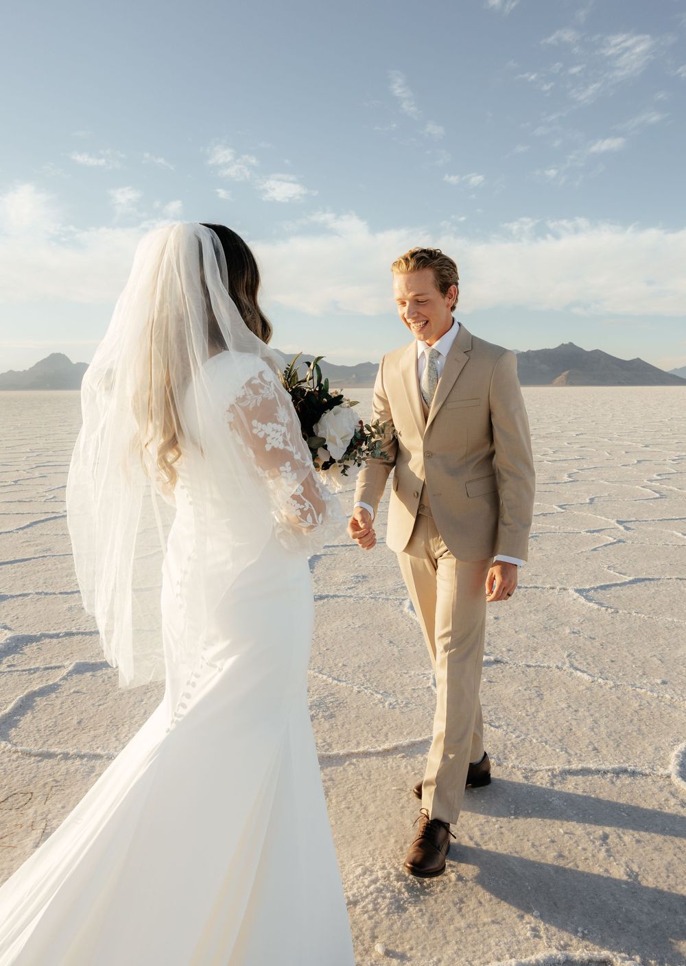Groom in beige suit holds bride's hand while walking on the Bonneville Salt Flats with mountains behind.