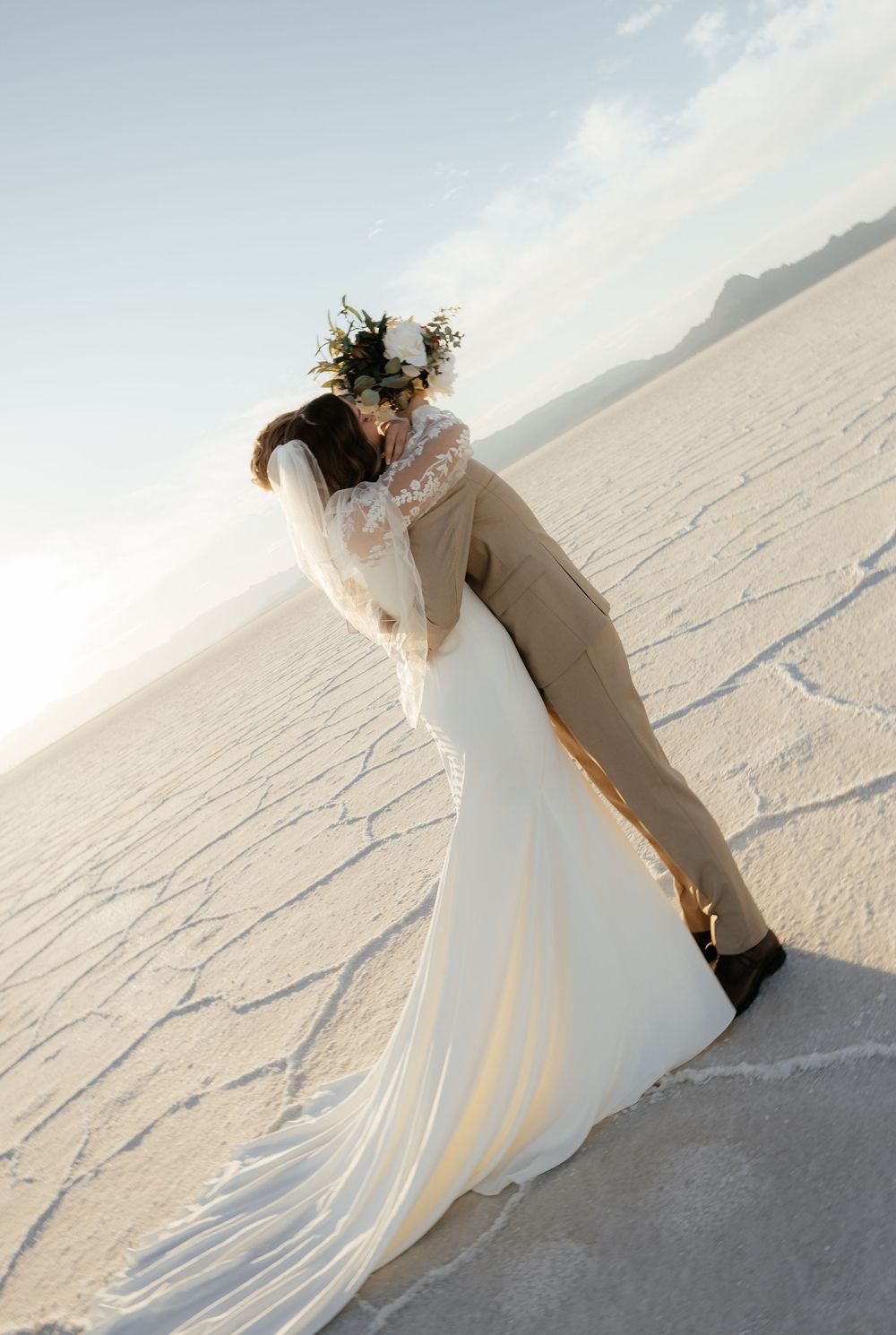 Bride and groom share a dip kiss on the white salt flats, bride wearing floral crown and lace gown.