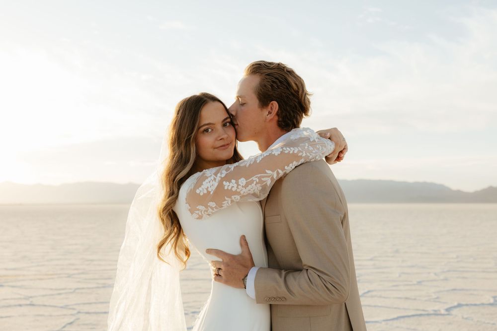 Groom kisses bride on the cheek as she smiles on the glowing white Bonneville Salt Flats.