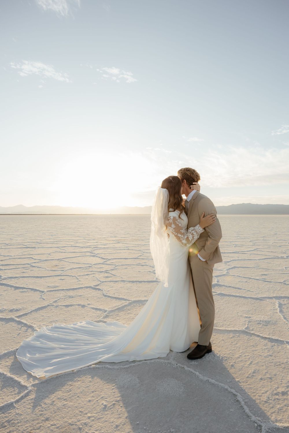 Bride and groom kiss at sunset on the Bonneville Salt Flats, veil trailing across the cracked earth.