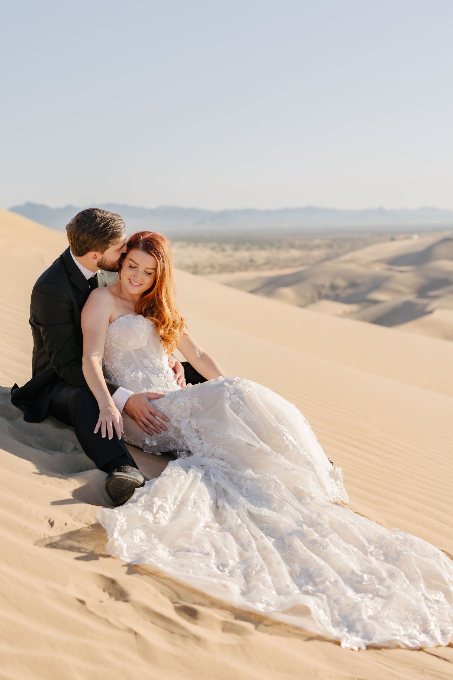 A couple in wedding attire sitting together on golden sand dunes during sunset.