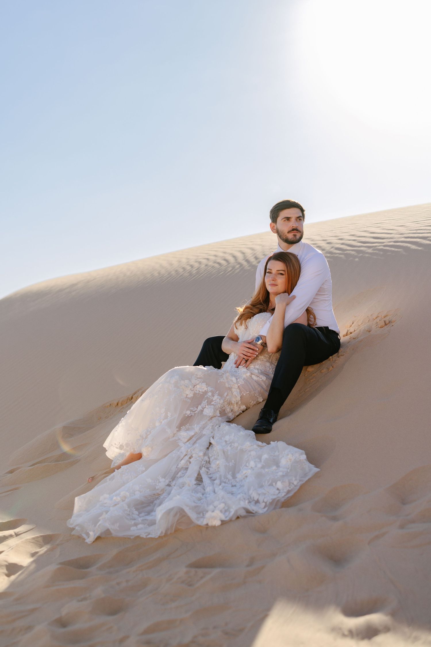 A couple in wedding attire sitting on a steep desert sand dune.