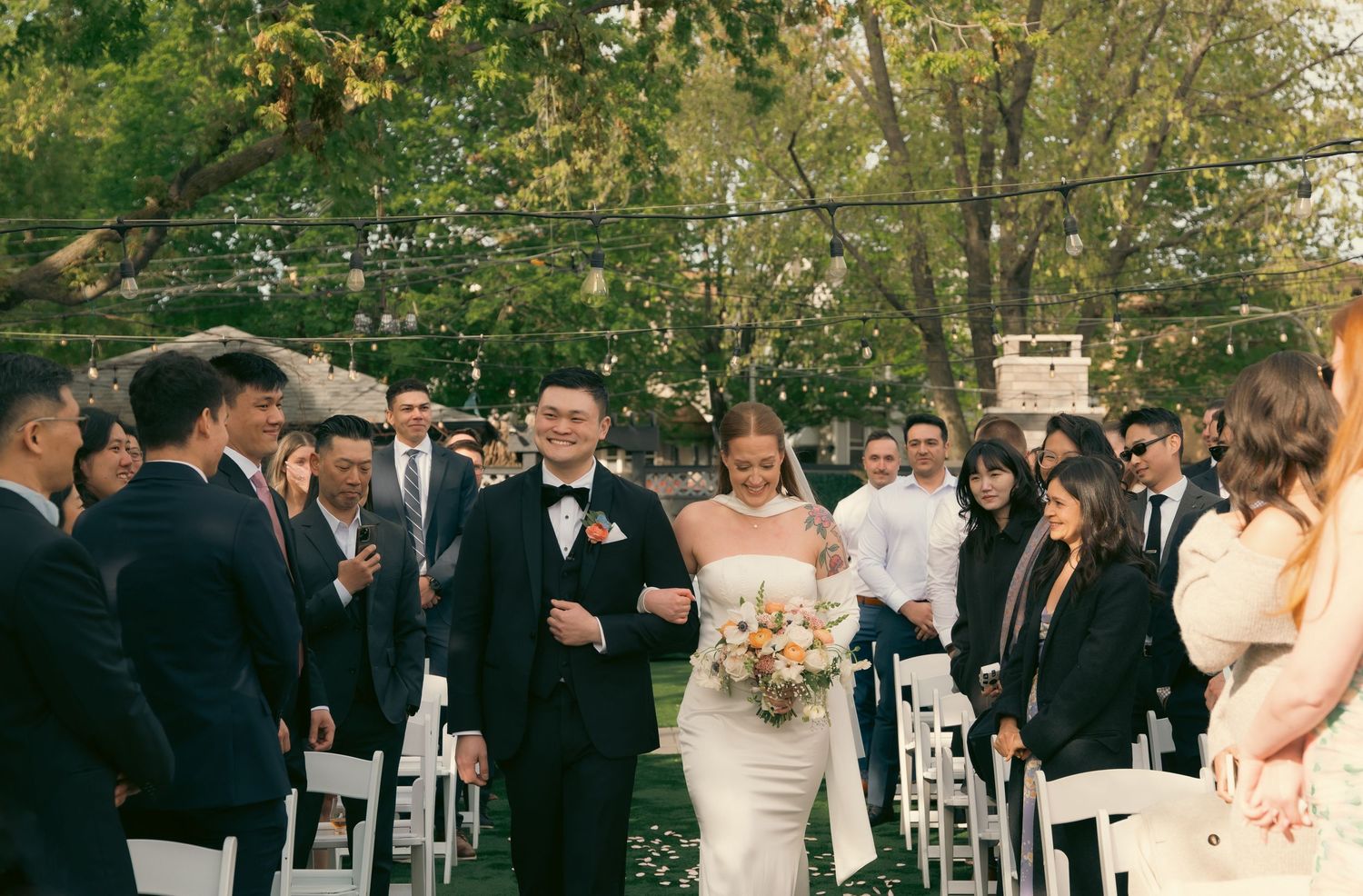 A bride in a white lace gown celebrates with friends in black dresses during a garden reception.