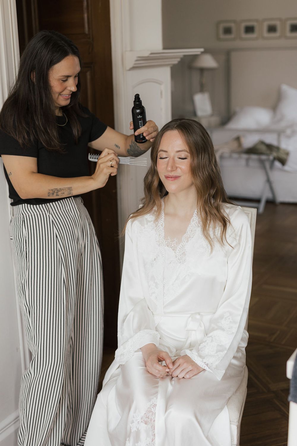 Wedding preparation scene showing person in white robe getting hair styled while seated in elegant room.